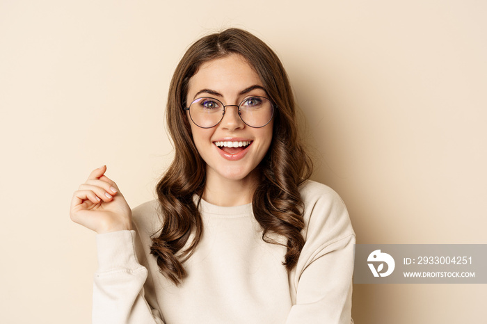 Close up portrait of stylish brunette woman in glasses, laughing and smiling, posing in eyewear against beige background