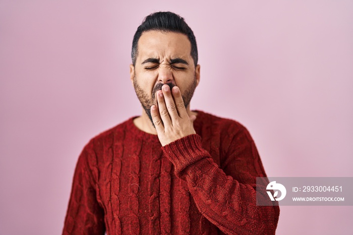 Young hispanic man with beard wearing casual sweater over pink background bored yawning tired covering mouth with hand. restless and sleepiness.