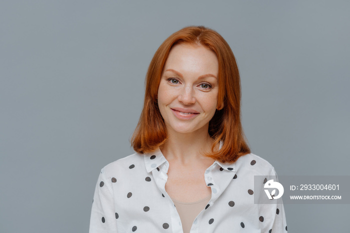 Portrait of attractive ginger European woman smiles gently at camera, wears makeup, dressed in polka dot blouse, looks directly at camera, models against grey background, thinks about changing job