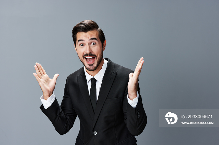 The man business in the suit is surprised and smiling a smile with his teeth raising his hands up against the gray background