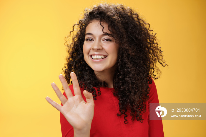 Charming friendly and self-assured attractive curly woman waving cute with palm to say hi or hello smiling broadly greeting man trying flirt in party posing joyful over yellow background