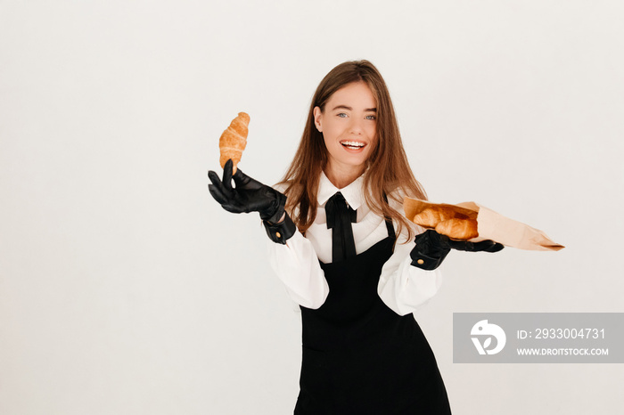 Laughing waitress in a uniform and black gloves posing with fresh croissants, ready to have lunch, looking at the camera on a white background