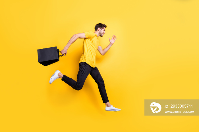 Full size photo of handsome guy jumping high holding packages speed rushing sale shopping wear casual t-shirt pants isolated yellow color background