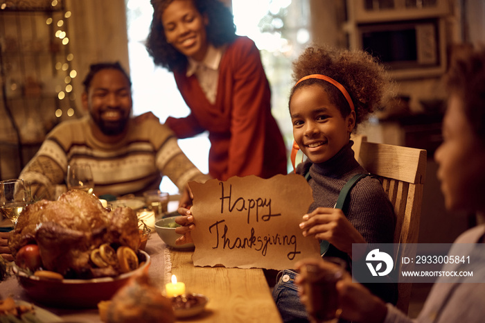 Happy black girl holding Happy Thanksgiving sign while having dinner with her family at dining table and looking at camera.