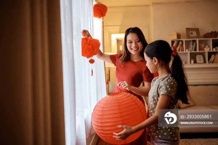 Happy Chinese mother and her daughter using lanterns while decorating home for Lunar New Year celebration.