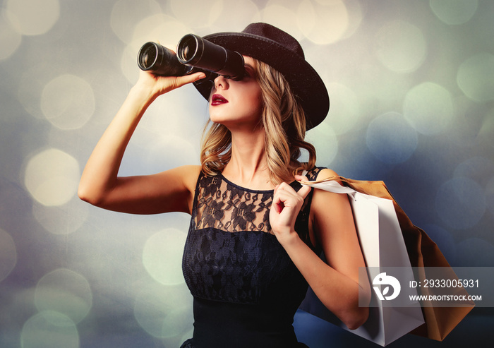 Portrait of a young style girl in black dress and hat with shopping bags and binoculars on gray background with bokeh