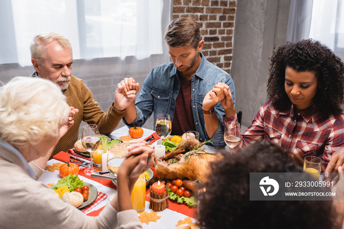 selective focus of multiethnic family with closed eyes holding hands during dinner on thanksgiving holiday