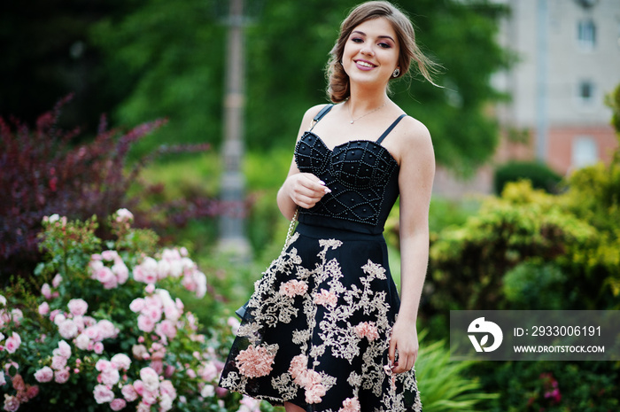 Portrait of a gorgeous young girl in black floral dress walking on the pavement with leather bag in the park on a prom day.