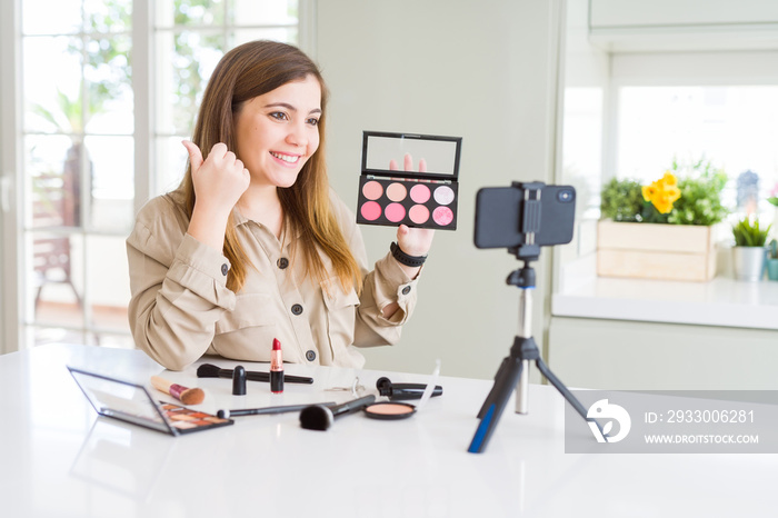 Beautiful young influencer woman recording make up video tutorial pointing and showing with thumb up to the side with happy face smiling