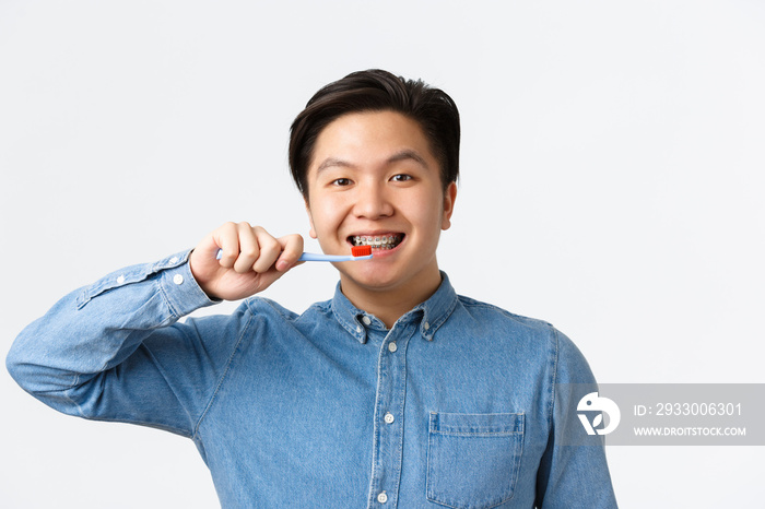 Orthodontics, dental care and hygiene concept. Close-up of friendly-looking smiling asian man brushing teeth with braces, holding toothbrush, standing white background