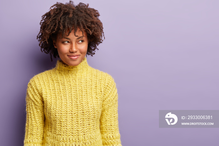 Headshot of curious satisfied dark skinned woman with curly haircut, looks away with intention or intriguing plan, wears yellow clothes, has something in mind, poses against purple wall, blank space