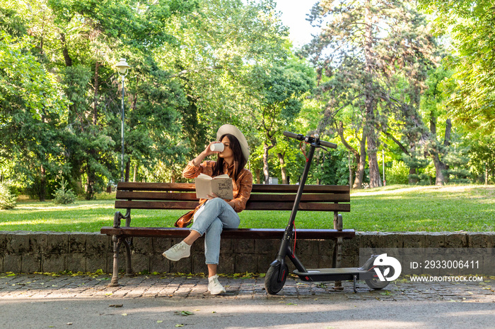 A young girl is sitting on a park bench, reading book and drinking coffee