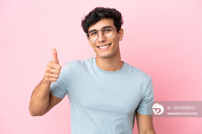 Young Argentinian man isolated on pink background With glasses and with thumb up