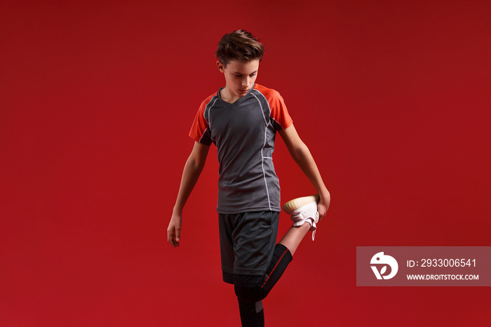 Young sportsman. A teenage boy is engaged in fitness, he is looking aside while stretching his legs. Isolated on red background. Sport, training, active lifestyle concept. Horizontal shot