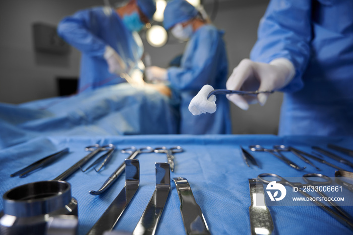 Close up of doctor hand holding forceps with tampon while surgeon and assistant performing plastic surgery on blurred background. Medical team doing cosmetic surgery in operating room at hospital.