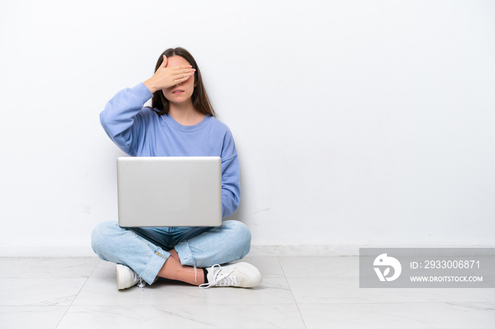 Young caucasian woman with laptop sitting on the floor isolated on white background covering eyes by hands. Do not want to see something