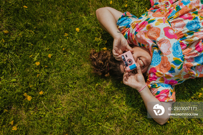 woman lies on grass taking a picture with a pink camera