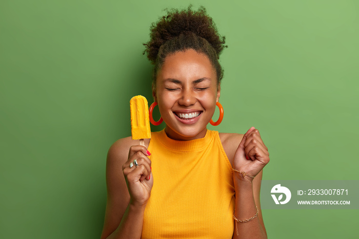 Happy dark skinned millennial girl eats yellow ice cream of mango flavor, clenches fist with anticipation of something awesome happen, has toothy smile, wears bright summer clothes, isolated on green