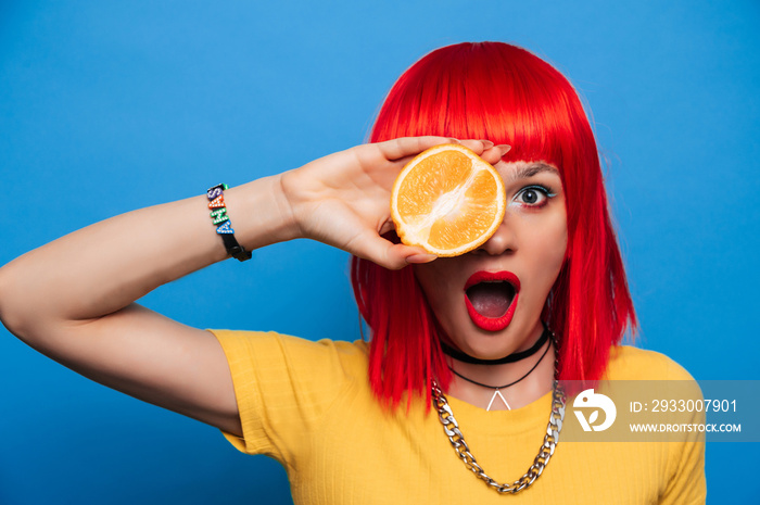 A young beautiful funny model with a slice of orange near her eyes, in the background with makeup, hairstyle. Bright, funny summer photo. Bright background.