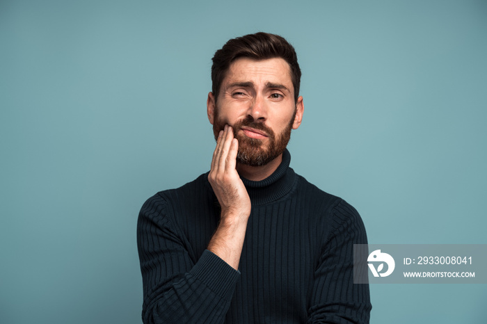 Dental problems. Portrait of unhealthy man pressing sore cheek, suffering acute toothache, periodontal disease, cavities or jaw pain. Indoor studio shot isolated on blue background