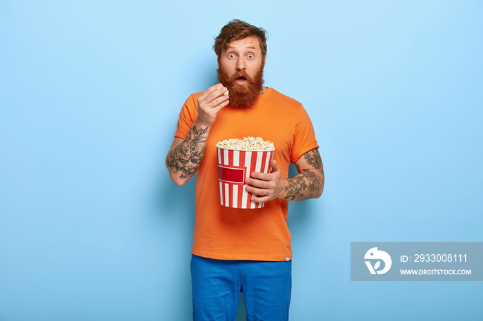 Ginger shocked man eats popcorn, watches horror film alone at night, has impressive look, wears orange t shirt, watches TV, isolated on blue background. Evening entertainment during spare time