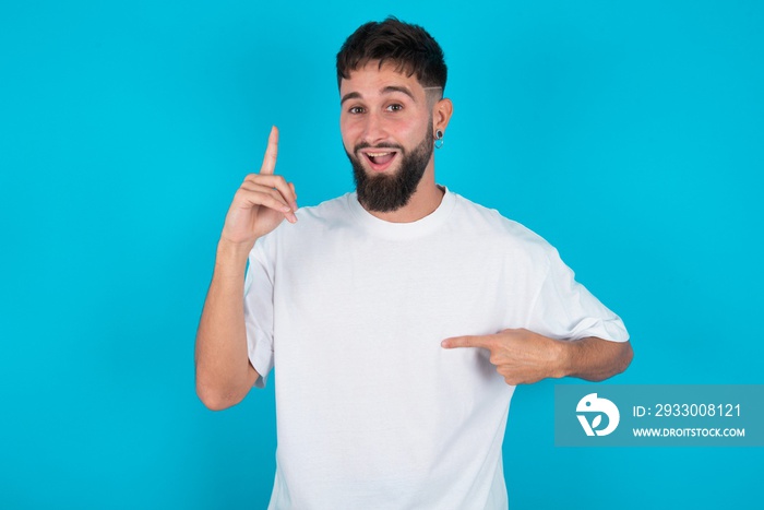 bearded caucasian man wearing white T-shirt over blue background says: wow how exciting it is, has amazed expression, indicates something. One hand on her chest and pointing with other hand.