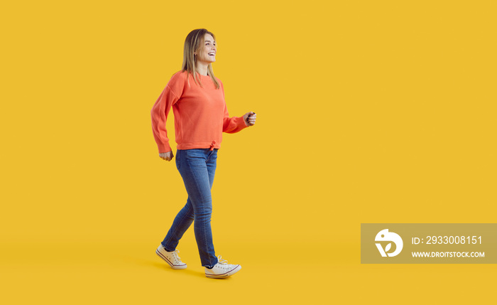 Full body studio shot of a cheerful smiling girl wearing orange sweatshirt and blue jeans. Happy young woman in a comfortable casual outfit walking forward against a vibrant yellow colour background