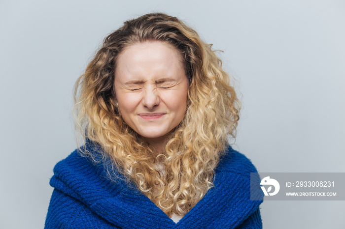 Horizontal shot of lovely young woman keeps eyes shut as anticipates something, going to recieve present, doesn`t open eyes, wears knitted sweater, isolated over light blue studio background