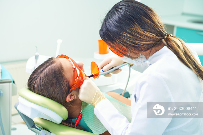 Girl child at the doctor. Dentist places a filling on a tooth with dental polymerization lamp in oral cavity. over clinic background