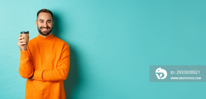 Handsome smiling man taking break, drinking coffee from takeaway, standing over blue background