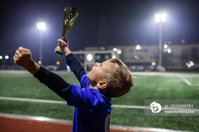 Young soccer player in blue jersey with ten number raising a trophy after the winning goal in the football competition, illuminated stadium, sport, winner and success. Dreams come true