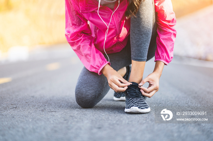 Close up athlete Asian girl running exercise fitness healthy lifestyle resting tying shoe lace on road listening music earphones wearing jogging clothes, morning sunrise outdoor urban road trail