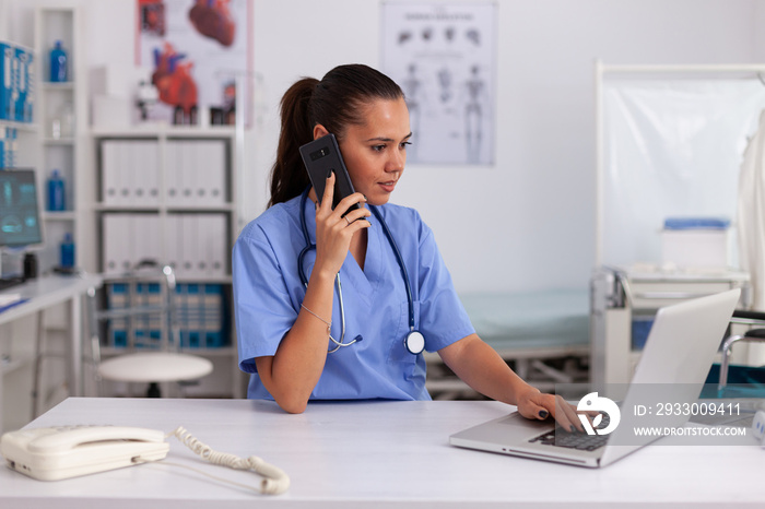 Medical practitioner using telephone and laptop in hospital office wearing blue uniform. Health care physician sitting at desk using computer in modern clinic looking at monitor.