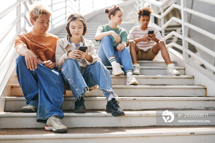 Airy full length shot of diverse teenagers using smartphones outdoors while sitting on metal stairs in sunlight