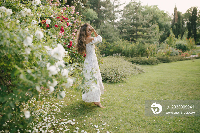 Young beautiful woman with long curly hair wearing white dress relaxing in blooming rose garden.