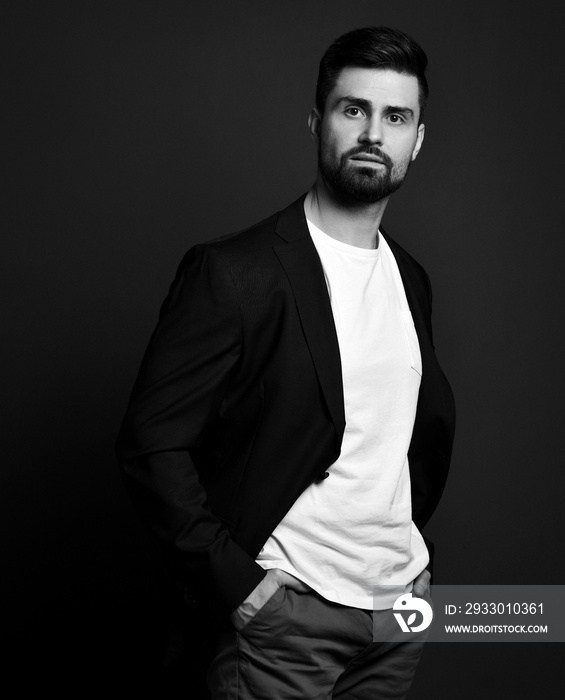 Handsome brutal man in stylish casual clothes and jacket standing with hands in pockets and looking at camera over dark background. Stylish business casual style for men