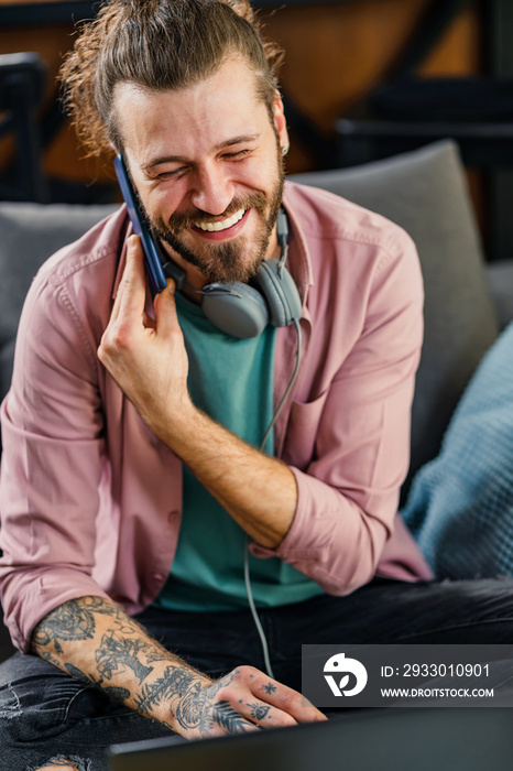 Handsome young hipster man working on a laptop computer at home, using a smartphone.