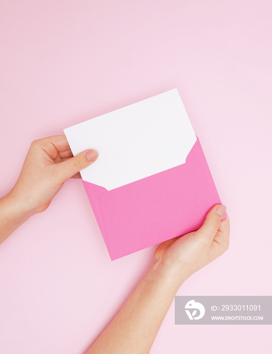 Female hands with envelope and blank card. Woman holding pink envelope square invitation card mockup on pink background. Top view with copy space. Template for branding and advertising