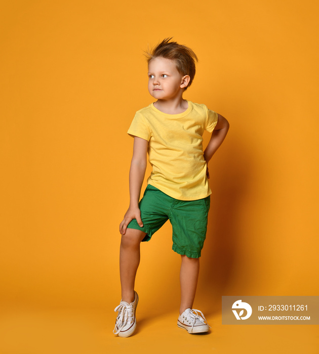 Young boy kid in yellow t-shirt and green shorts stands with hand on his knee on yellow background