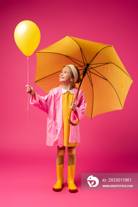 full length of cheerful girl in raincoat holding balloon and yellow umbrella on crimson