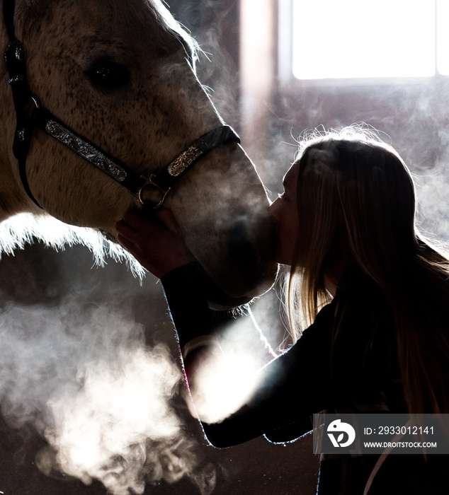 Low key image. horse and girl in barn during cold winter day, steamy breath visible back lit from window