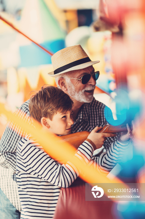 Grandfather and grandson having fun and spending good quality time together in amusement park. Kid shooting with air gun while grandpa helps him to win the prize.