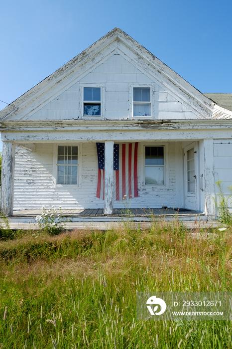 Vertical image of an old abandoned farmhouse with an american flag on the large empty abandoned veranda porch.