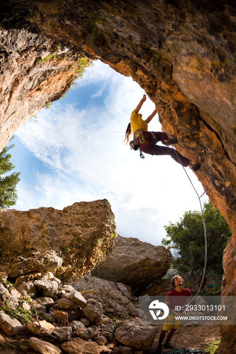 The girl climbs the rock in the shape of an arch, A man is belaying a climbing partner.