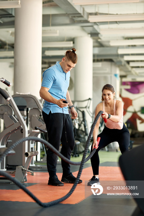 Happy athletic woman practicing with battle ropes during cross training with her coach in a gym.