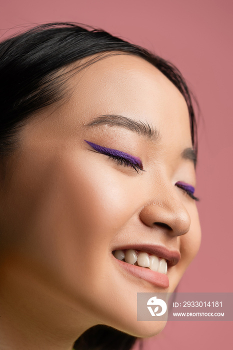 close up view of young asian woman with makeup and blue eyeliner smiling with closed eyes isolated on pink.