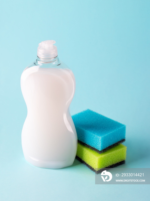 White dishwashing liquid in a plastic bottle and two foam sponges. Purity and household chemicals. Kitchen detergent on a pastel blue background.