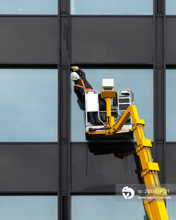 Male window cleaner cleaning glass windows on modern building high in the air on a lift platform. Worker polishing glass high in the air