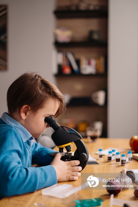 Distance online education, internet learning. First grade boy studying at home using microscope, making notes, biology online lesson