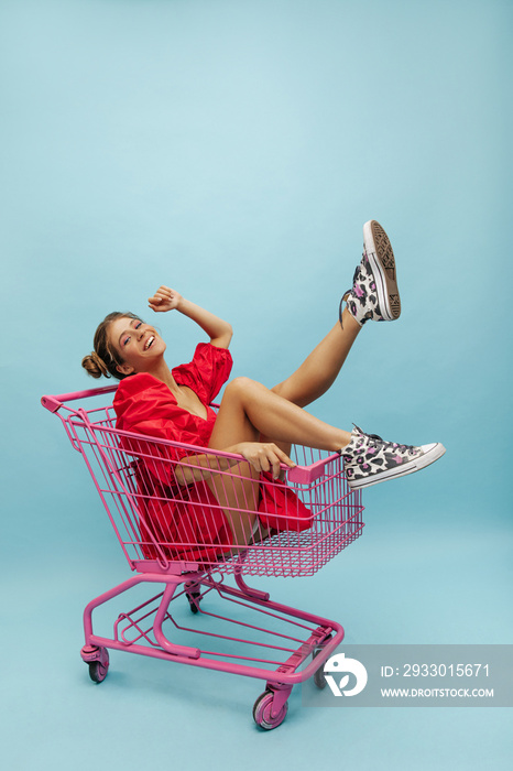 Young cool caucasian smiling hipster woman smiling while sitting in trolley on blue background. Tanned model with gathered blonde hair in bright red outfit. Shopping concept